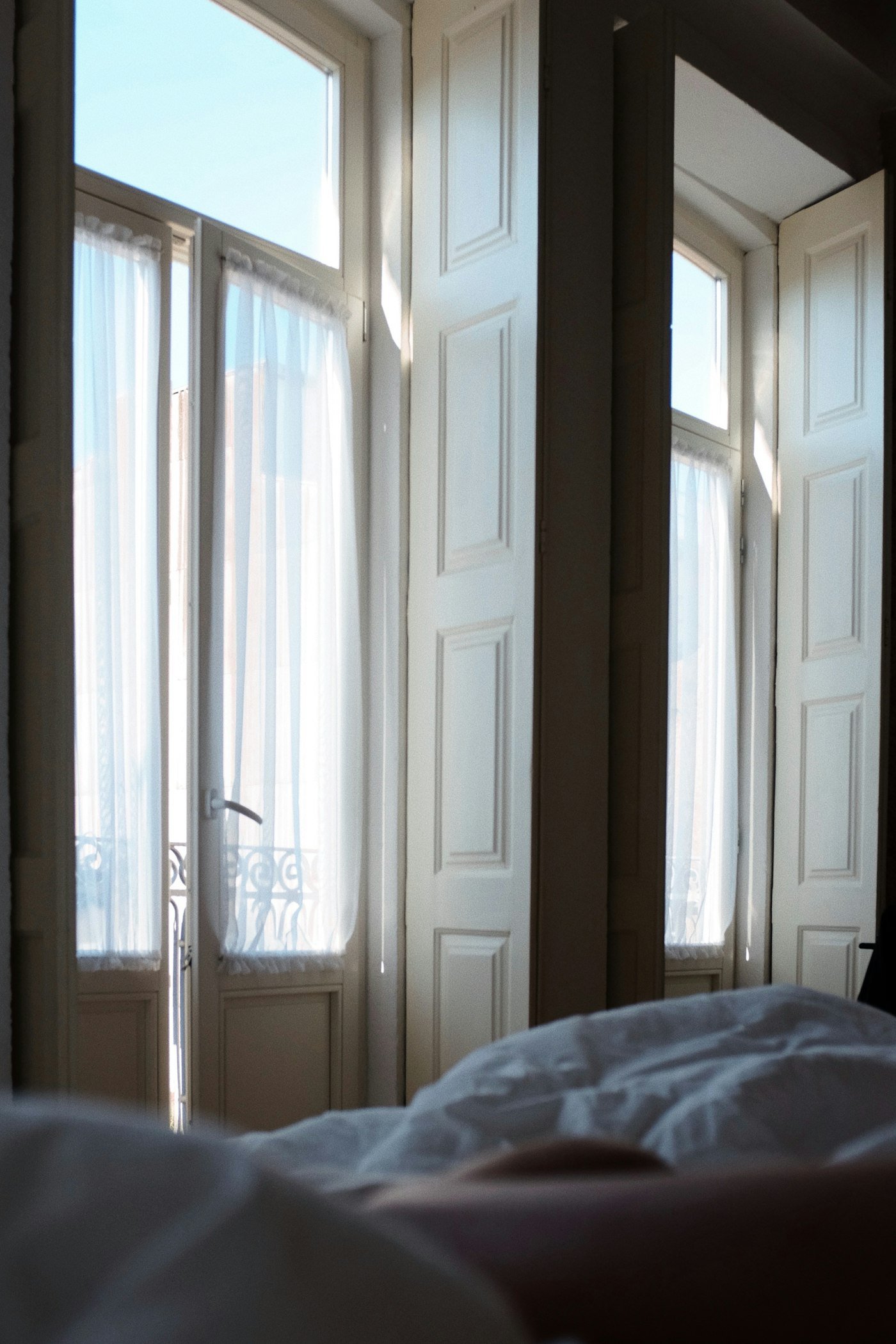 Soft morning light pouring through tall windows over an unmade bed